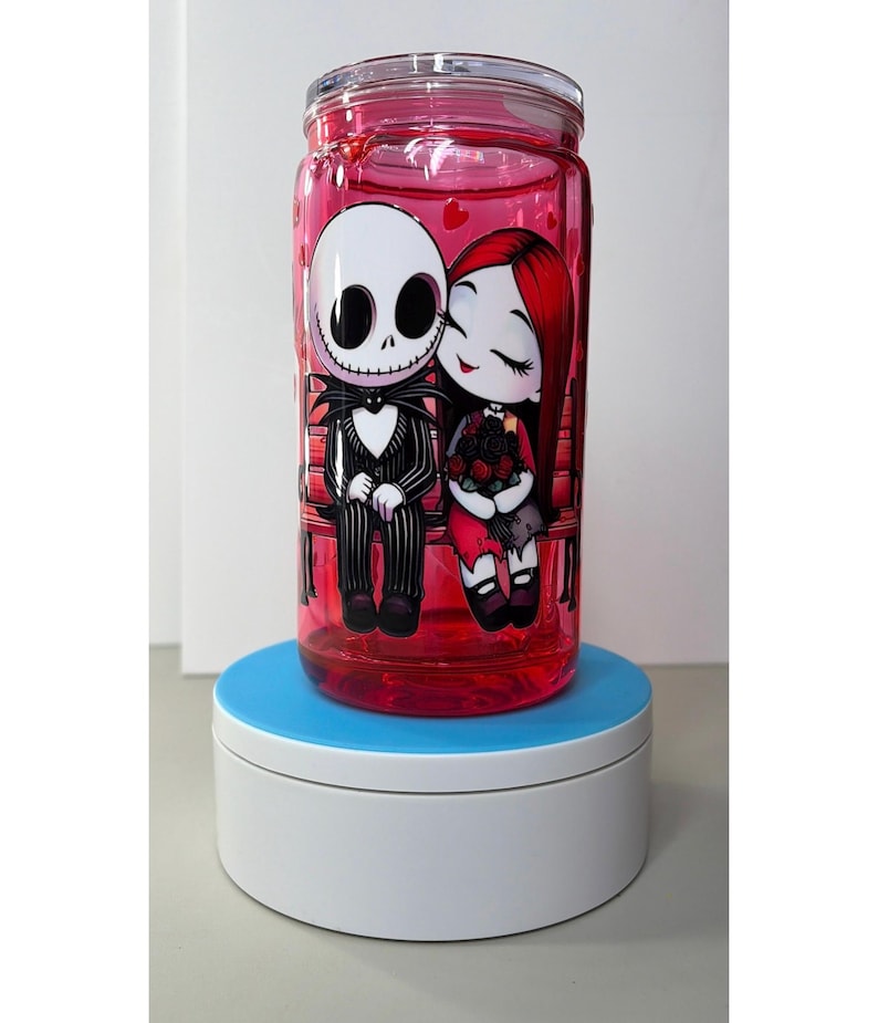 Op de afbeelding: Een heldere, cilindrische beker met een rode vloeistof erin en een schroefdeksel. De beker heeft een cartoonillustratie van Jack Skellington en Sally uit The Nightmare Before Christmas. De personages zitten op een bank, omringd door harten.