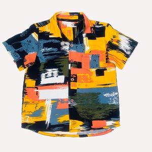 Può includere: Camicia a maniche corte con un vivace design astratto. La camicia presenta una base gialla con motivi a pennellate in nero, bianco, blu e arancione. Ha una tasca sul petto e un colletto classico.