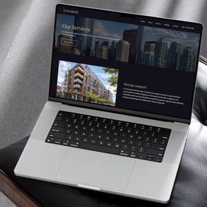 May include: A silver laptop displaying a website with the text "LUNA MEAVE" at the top. The website features images of buildings and text about services and buying support. The laptop is on a black surface.
