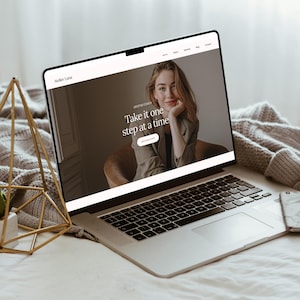 May include: A silver laptop showing a website with the text "Take it one step at a time." The website features a photo of a woman and the words "Lifestyle Coach." A gold geometric decoration and a smartphone are also visible.