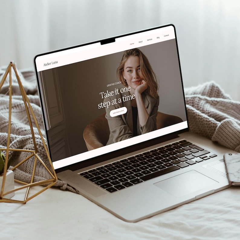 May include: A silver laptop showing a website with the text "Take it one step at a time." The website features a photo of a woman and the words "Lifestyle Coach." A gold geometric decoration and a smartphone are also visible.