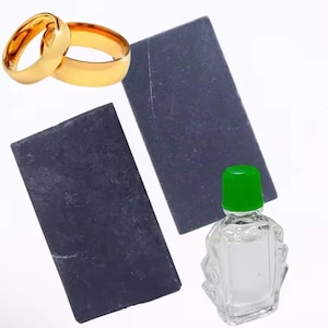 May include: Two gold wedding bands, two rectangular black stones, and a small clear glass bottle with a green cap are displayed against a white background. The bottle is filled with a clear liquid.