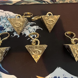 May include: Five gold-colored, pyramid-shaped pendants with an eye symbol are displayed. Each pendant has a loop at the top for attachment. The pendants are on a patterned surface with a wave design.