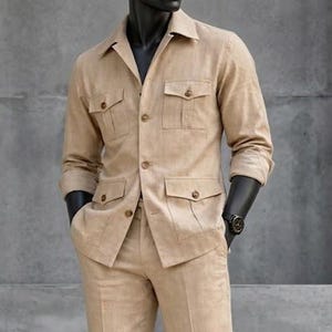 May include: A tan linen suit on a mannequin. The suit includes a long-sleeved jacket with four front pockets and matching flared trousers. The mannequin wears a black watch and black shoes. The background is a gray concrete wall.