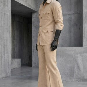 May include: A tan linen suit on a mannequin. The suit includes a collared jacket with four pockets and matching flared pants. The mannequin wears a black watch and black gloves. The suit is a neutral color and has a classic design.