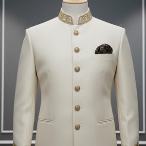 May include: Cream-coloured formal suit with a mandarin collar and gold embroidered details. The suit features ornate gold buttons and cuff embroidery. A patterned pocket square adds a touch of elegance. The suit is displayed on a mannequin.