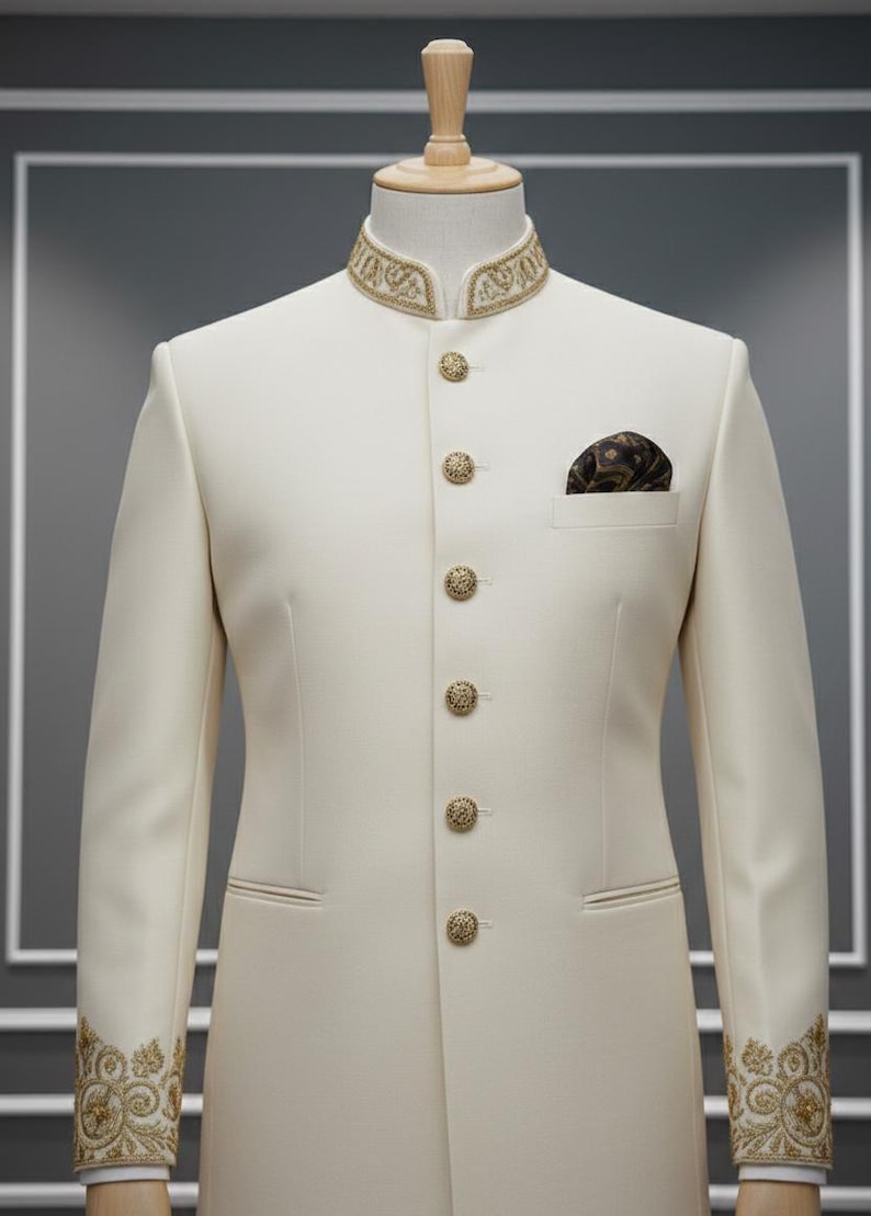 May include: Cream-coloured formal suit with a mandarin collar and gold embroidered details. The suit features ornate gold buttons and cuff embroidery. A patterned pocket square adds a touch of elegance. The suit is displayed on a mannequin.