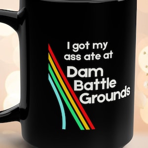 Könnte beinhalten: Schwarze Keramik-Tasse mit großem Henkel. Die Tasse zeigt den Text "I got my ass ate at Dam Battle Grounds" in Weiß, mit einer farbenfrohen Grafik aus roten, gelben, grünen und blauen Linien.