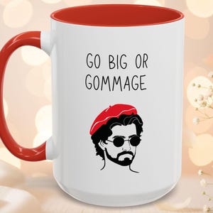May include: White ceramic mug with a red interior and handle. The mug features the text "GO BIG OR GOMMAGE" and an illustration of a man in a red beret and sunglasses. The mug is a humorous gift idea.