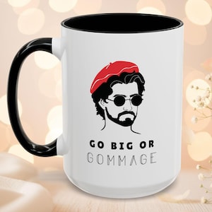 May include: White ceramic mug with a black handle and interior. The mug features a graphic of a man in a red beret and sunglasses, with the text "GO BIG OR GOMMAGE" in black. Suitable for tea, coffee, or other hot drinks.