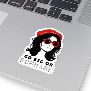 May include: A white sticker featuring a stylized illustration of a woman with black hair, red sunglasses, and a red beret. The sticker has the text "GO BIG OR GOMMAGE" in a black font.