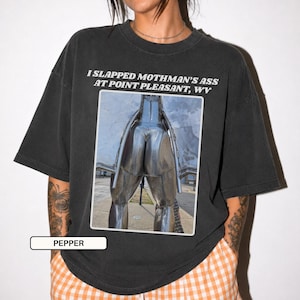 May include: A charcoal gray short-sleeve t-shirt featuring a rectangular graphic print of a metallic sculpture. The text above the image reads, I SLAPPED MOTHMAN'S ASS AT POINT PLEASANT, WV. The shirt has a crew neckline and a relaxed fit.