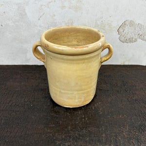 Op de afbeelding: Een cilindrische keramische pot met een glanzend, lichtgeel glazuur en twee kleine, gebogen handvatten aan weerszijden. De pot heeft een dikke, afgeronde rand en een gladde, enigszins onregelmatige textuur met tekenen van ouderdom.