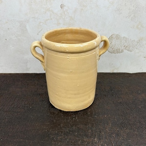 Op de afbeelding: Een cilindrische keramische pot met een gladde, lichtgele geglazuurde afwerking. De pot is voorzien van twee kleine, gebogen handvatten aan weerszijden bij de rand, een brede ronde opening en een licht taps toelopende onderkant.