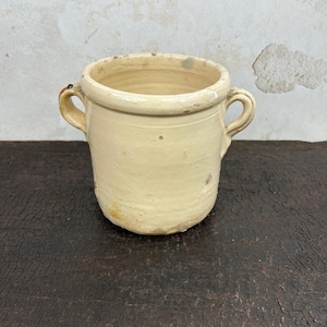 Op de afbeelding: Een crèmekleurige keramische pot met een gladde, geglazuurde afwerking en twee handvatten aan de zijkant. De pot heeft een brede, open bovenkant en een cilindervormig lichaam met subtiele onregelmatigheden en een licht verweerd uiterlijk.