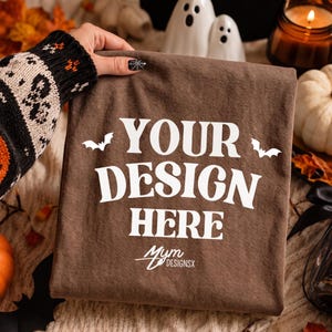 May include: A brown t-shirt featuring the white text "YOUR DESIGN HERE" in a bold, stylised font, flanked by two small white bat silhouettes. Below the text, the logo "Mym DESIGNSX" is printed in white. The fabric has a smooth, matte texture.