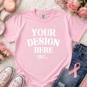 May include: A light pink short-sleeve t-shirt featuring the white text "YOUR DESIGN HERE" printed in the center. The shirt has a crew neckline and a Comfort Colors brand label on the inside collar. The design also includes the text "MYM DESIGNSX".