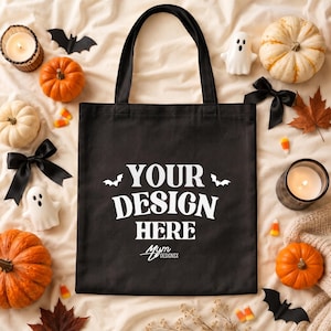 Puede incluir: Un bolso de tela negro con dos asas para el hombro que presenta el texto blanco "YOUR DESIGN HERE" centrado en la parte delantera, flanqueado por dos pequeñas siluetas de murciélagos blancos, con el logo "MYM DESIGNSX" impreso en blanco.