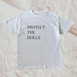 May include: White t-shirt with the text "PROTECT THE DOLLS" in black, centered on the front. The shirt has a crew neck and short sleeves. The fabric appears to be cotton or a cotton blend.