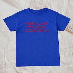 Op de afbeelding: Een koningsblauw t-shirt met de rode tekst "KISS ALL THE TIME. DISCO, OCCASIONALLY." Het shirt met korte mouwen is gemaakt van een zacht materiaal en is geschikt voor vrijetijdskleding. Het shirt wordt gepresenteerd op een lichtbruin, getextureerd oppervlak.