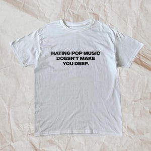 Könnte beinhalten: Weißes T-Shirt mit dem schwarzen Text "HATING POP MUSIC DOESN'T MAKE YOU DEEP" mittig auf der Vorderseite. Das Shirt hat kurze Ärmel und einen Rundhalsausschnitt. Der Hintergrund ist eine hellbraune, strukturierte Oberfläche.