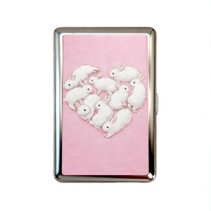 K&ouml;nnte beinhalten: Ein rechteckiges Metallgeh&auml;use mit einem polierten silbernen Rahmen und einer hellrosa Frontplatte. Die Platte zeigt eine grafische Illustration von neun wei&szlig;en Kaninchen mit rosa get&ouml;nten Ohren, die in Herzform auf dem rosa Hintergrund angeordnet sind.