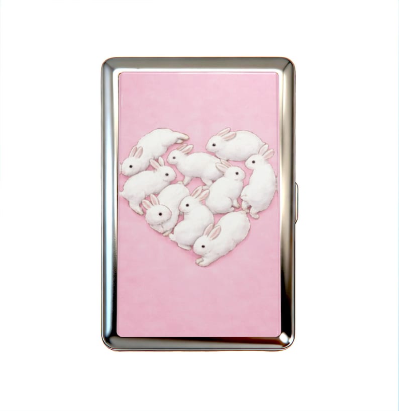 K&ouml;nnte beinhalten: Ein rechteckiges Metallgeh&auml;use mit einem polierten silbernen Rahmen und einer hellrosa Frontplatte. Die Platte zeigt eine grafische Illustration von neun wei&szlig;en Kaninchen mit rosa get&ouml;nten Ohren, die in Herzform auf dem rosa Hintergrund angeordnet sind.