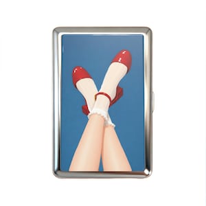 K&ouml;nnte beinhalten: Ein rechteckiges Zigarettenetui aus Metall mit einer gl&auml;nzenden Illustration von &uuml;berkreuzten Beinen, die wei&szlig;e Socken mit R&uuml;schen und gl&auml;nzende rote Mary-Jane-Schuhe tragen, vor einem einfarbigen blauen Hintergrund. Polierter silberner Rahmen.
