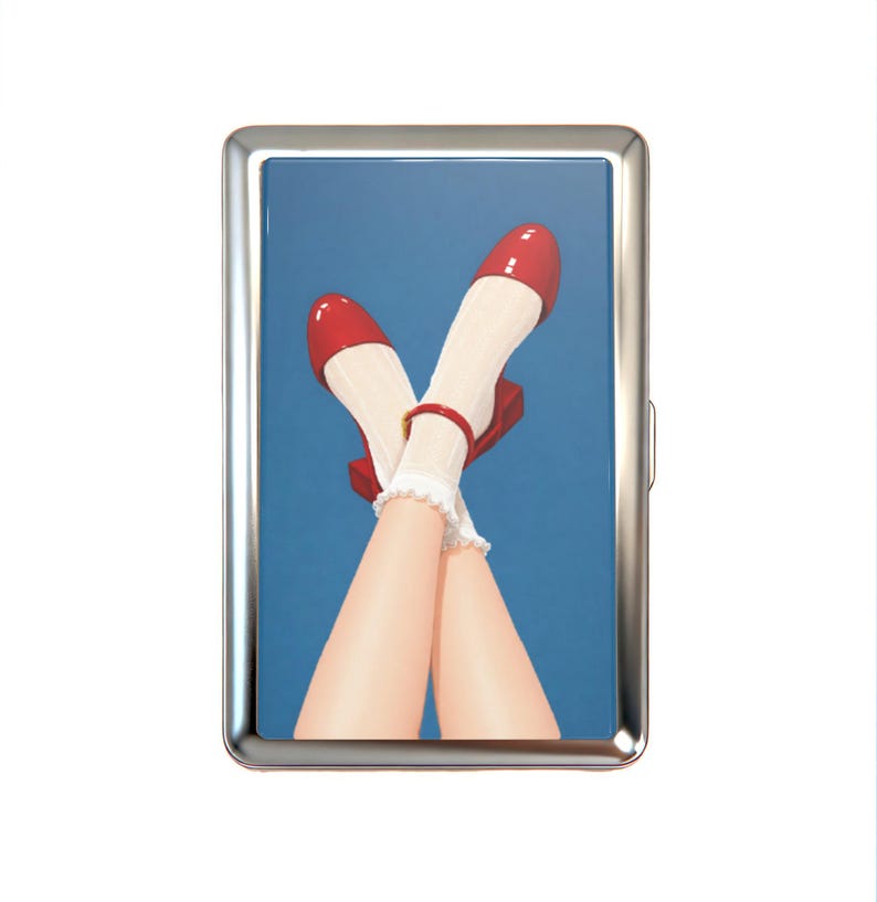 K&ouml;nnte beinhalten: Ein rechteckiges Zigarettenetui aus Metall mit einer gl&auml;nzenden Illustration von &uuml;berkreuzten Beinen, die wei&szlig;e Socken mit R&uuml;schen und gl&auml;nzende rote Mary-Jane-Schuhe tragen, vor einem einfarbigen blauen Hintergrund. Polierter silberner Rahmen.