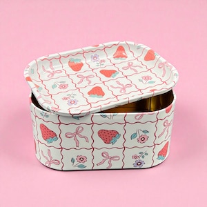 May include: A rectangular tin box with a matching lid, decorated with a repeating pattern of strawberries, bows, and flowers. The box and lid are white with red and pink accents, set against a pink background.