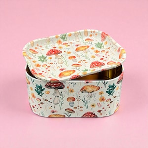 May include: A rectangular metal storage tin with a lid, featuring a whimsical mushroom and floral pattern. The design includes red, orange, and brown mushrooms, along with green foliage and yellow flowers, set against a cream background. The tin is open, revealing a gold interior.