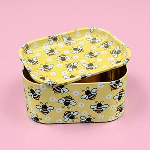 May include: A yellow metal storage tin with a matching lid. The tin and lid are decorated with a repeating pattern of cartoon bees and white daisies. The background is a solid pink.