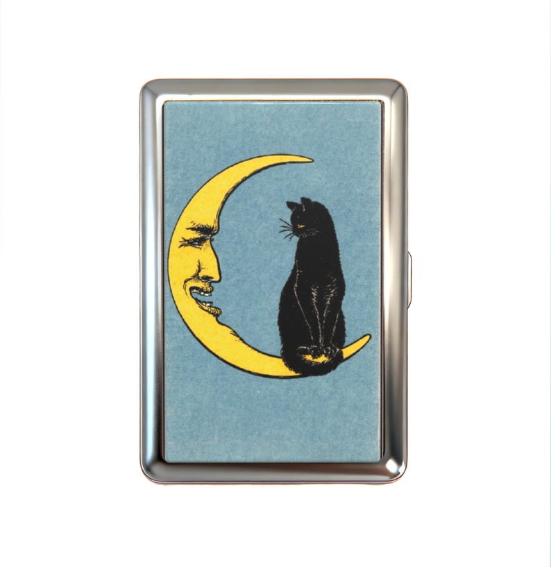 K&ouml;nnte beinhalten: Ein rechteckiges Etui aus silberfarbenem Metall mit einer zentralen Illustration auf hellblauem Hintergrund. Das Bild zeigt eine gelbe Mondsichel mit einem menschlichen Gesicht und eine schwarze Katze, die auf der unteren Mondsichel sitzt.