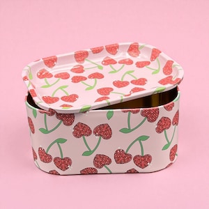 May include: A rectangular metal storage tin with a matching lid, both adorned with a repeating pattern of red cherries on a light pink background. The cherries have a checkered design and green stems. The lid is slightly ajar, revealing the interior.