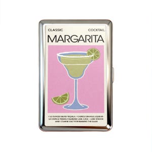K&ouml;nnte beinhalten: Ein rechteckiges Etui aus silbernem Metall mit einer Grafik eines Margarita-Cocktails auf rosa Hintergrund. Der Text lautet CLASSIC COCKTAIL MARGARITA und listet Zutaten wie 1 1/2 Unzen Silber-Tequila, 1 Unze Orangenlik&ouml;r und Limette auf.