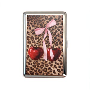 May include: A silver cigarette case featuring a leopard print background with a watercolor illustration of two red cherries tied with a pink ribbon. The case has a rectangular shape.