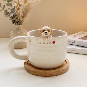 May include: Cream ceramic mug featuring a sculpted dog figurine on the rim. The mug has the word "Milano" and a red heart. The mug sits on a round wooden coaster. Decorative vase and lamp in the background.