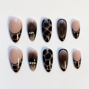 Könnte beinhalten: Ein Set aus zehn ovalen Kunstnägeln. Die Nägel haben ein Farbverlauf-Design, das von einer nackten Basis zu einer dunkelbraunen Spitze übergeht. Einige Nägel haben ein schwarzes, reptilienartiges Muster, andere kleine, dekorative Strasssteine.