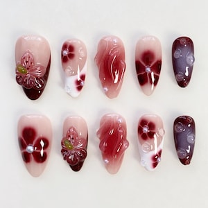 May include: A set of ten artificial nails in various designs. The nails feature a gradient of pink and burgundy hues, adorned with floral embellishments, pearls, and a glossy finish. Some nails have 3D flower accents.