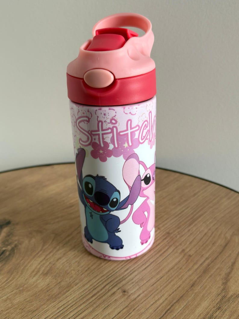 May include: A pink and red water bottle with the word "Stitch" in a pink font. The bottle features cartoon images of Stitch and Angel. It has a pink flip-top lid and is designed for hydration.