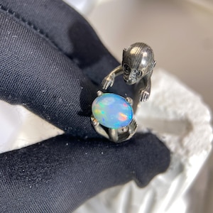 Könnte beinhalten: Ein silberner Ring mit einem Affen, der einen ovalen Opal-Edelstein hält. Der Opal zeigt ein Farbspiel mit Blau-, Grün- und Gelbtönen. Das Design des Rings ist detailliert und zeigt die Gesichtszüge des Affen und die Fassung des Edelsteins.