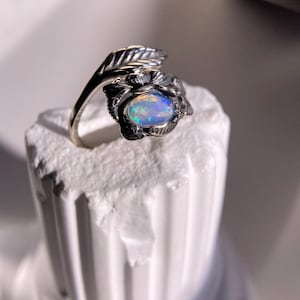 May include: A silver ring featuring an oval opal gemstone, set within a floral and leaf design. The opal displays a play of color with hues of blue, green, and yellow. The ring is displayed on a white, textured surface.