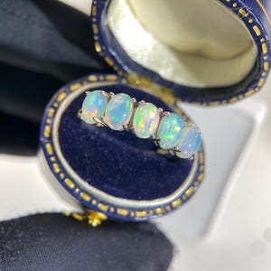 Peut inclure: Une bague en argent ornée de cinq opales ovales, chacune présentant un jeu de couleurs. La bague est présentée dans un écrin bleu avec une bordure dorée, sur fond sombre.