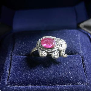 Peut inclure: Bague en argent ornée d'un motif éléphant et sertie d'une grosse pierre précieuse rose vif de forme ovale. La bague est présentée dans un écrin en velours bleu foncé.