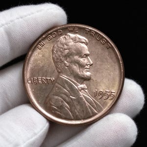 May include: A copper-coloured 1955 United States one-cent coin featuring the profile of Abraham Lincoln facing right. The coin displays the text IN GOD WE TRUST along the top edge, LIBERTY on the left side, and the year 1955 on the right side.