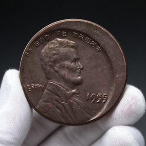 May include: A copper-colored 1955 Lincoln cent coin featuring a profile portrait of Abraham Lincoln facing right. The text IN GOD WE TRUST is arched above the head, LIBERTY is on the left, and the year 1955 is printed on the right side.
