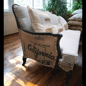Puede incluir: Un sofá de estilo vintage con estructura negra y laterales de arpillera. El asiento y el respaldo tienen tela a rayas y cojines con etiquetas "BEANS". Los laterales de arpillera muestran el texto "California" y otras marcas.