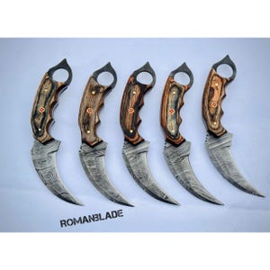 May include: Five karambit knives with curved blades and wooden handles. The handles are a mix of light and dark wood, with brass rivets. The blades have a patterned design. The text "ROMANBLADE" is at the bottom.