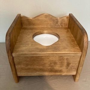 May include: A wooden potty chair with a medium brown stained finish, featuring a curved backrest and a smooth, flat seat surface. A circular cutout in the center of the seat reveals a white plastic basin insert, providing a functional design.