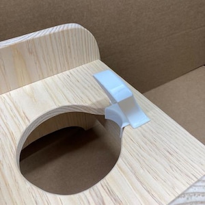 May include: A light-colored wooden stand featuring a circular cutout in the center and a white plastic clip attached to the edge of the opening. The wood grain is visible throughout the structure, which has a smooth, unfinished appearance.